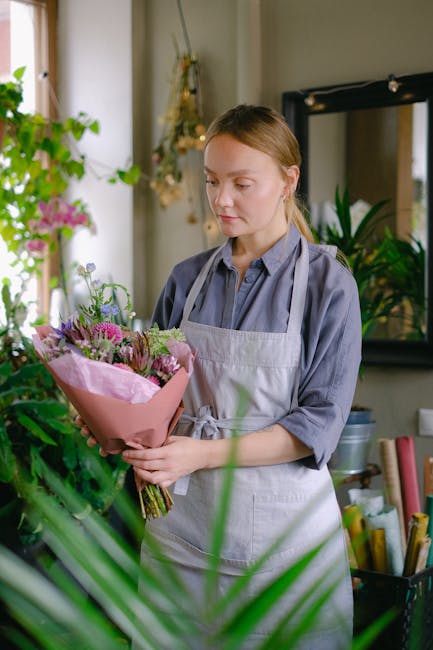 A young woman with light brown hair wearing a grey shirt and a white apron is holding a vibrant bouquet of fresh flowers wrapped in pink paper. The bouquet features a variety of blooms, including pink