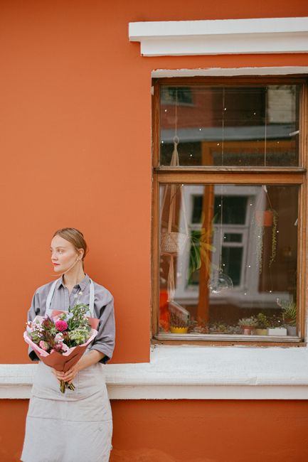 A young woman with fair skin and light brown hair styled in a loose, neat bun, holding a vibrant bouquet of fresh flowers wrapped in pink paper. The bouquet features an assortment of colorful blooms, 