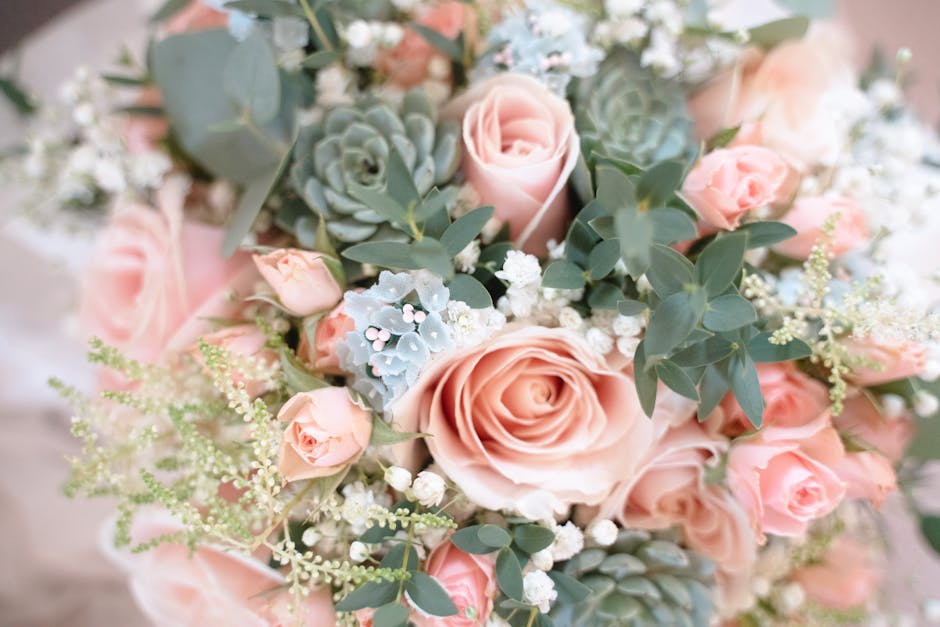 A close-up of a floral bouquet featuring large, soft pink roses, smaller matching pink spray roses, and delicate white baby's breath. Interspersed with silvery-green eucalyptus leaves and pale blue su