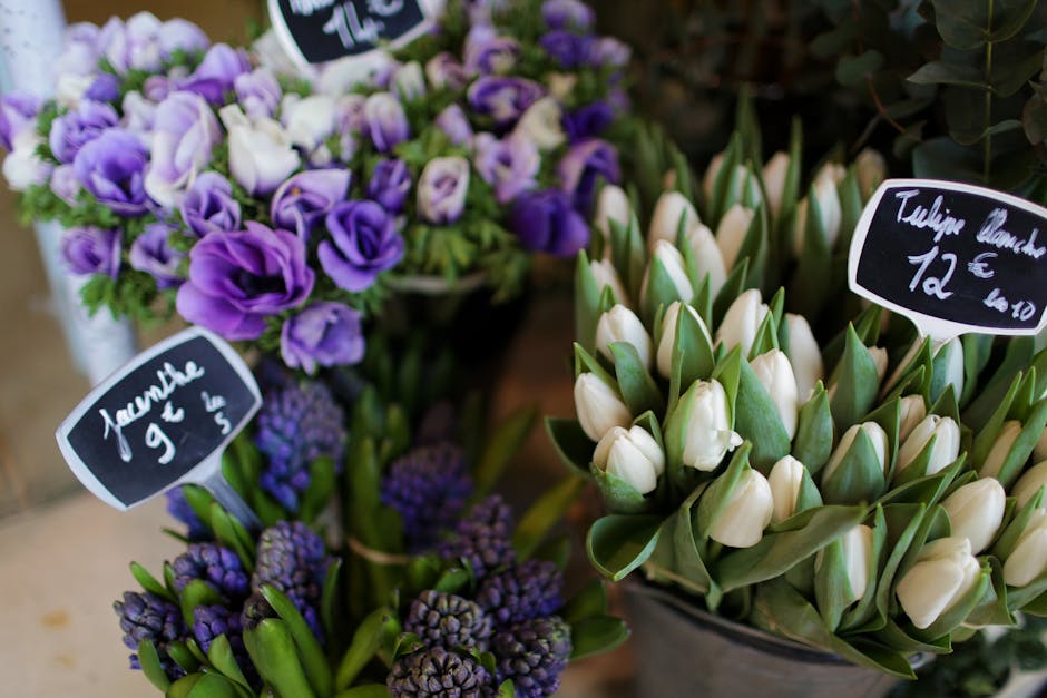 A display of fresh floral arrangements featuring purple and white flowers, including tulips and hyacinths. The tulips are tightly closed, creamy white with green edges, arranged in a neat bouquet with