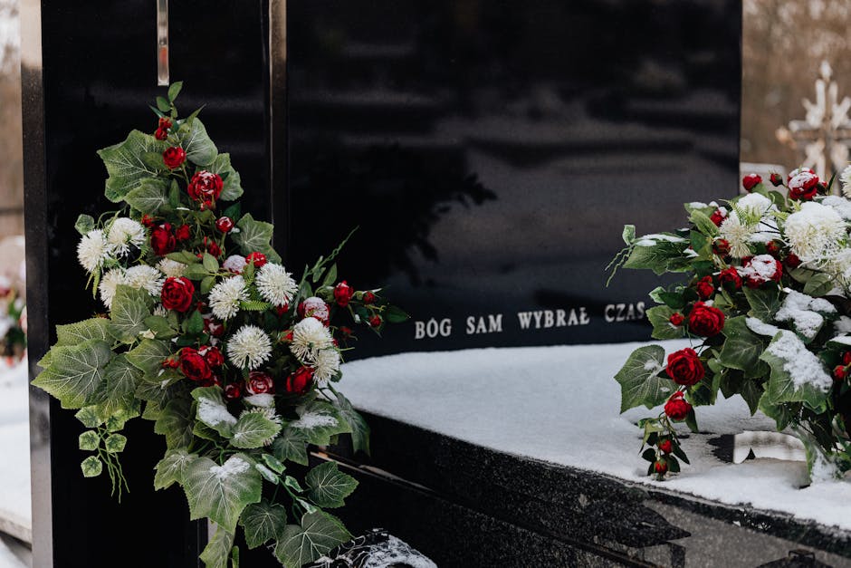 A polished black memorial gravestone adorned with floral tributes featuring red roses, white chrysanthemums, and green ivy leaves. The flowers are arranged in two bouquets placed on either side of the