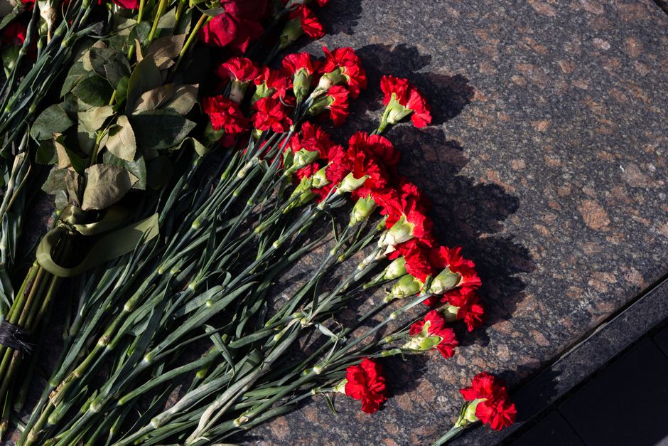 A bouquet of fresh red carnations with green stems and leaves, arranged loosely and laid on a dark granite surface outdoors. The carnations are wrapped at the base with a green ribbon, and some petals